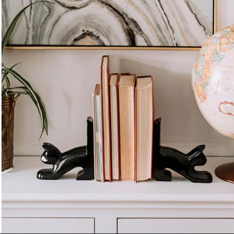 Black sculptural cat bookends holding a set of books on a white console table, with a potted plant on the left and a globe on the right, set against a wall with a large abstract agate print.