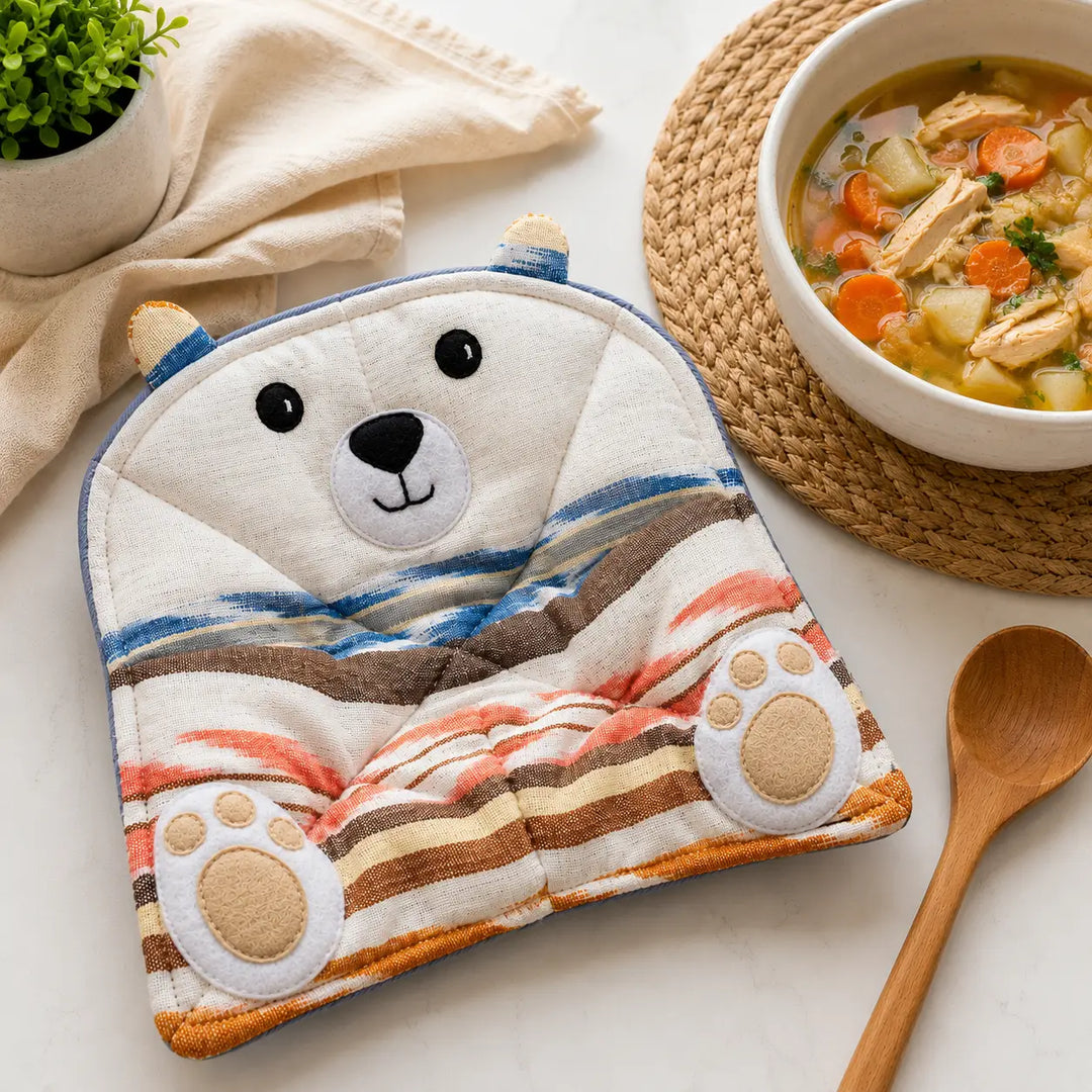 A quilted bear-shaped pot holder with a polar bear face and striped blanket design, styled on a tabletop next to a bowl of soup, a wooden spoon, and a small plant.