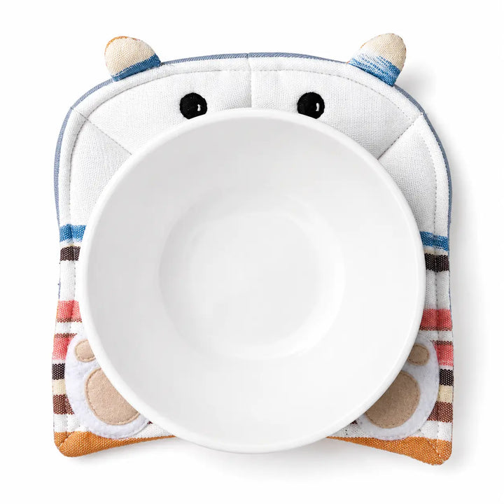 A white ceramic bowl centered on a plush, animal-face designed bowl cozy with embroidered eyes, ears, and striped details in blue, brown, pink, and beige, shown on a clean white background.