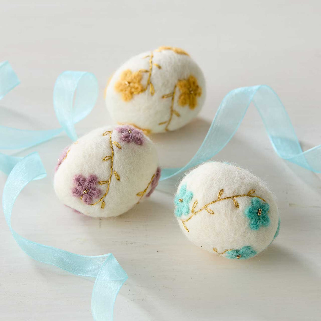 Three embroidered felt eggs with floral designs on a light surface with blue ribbons.