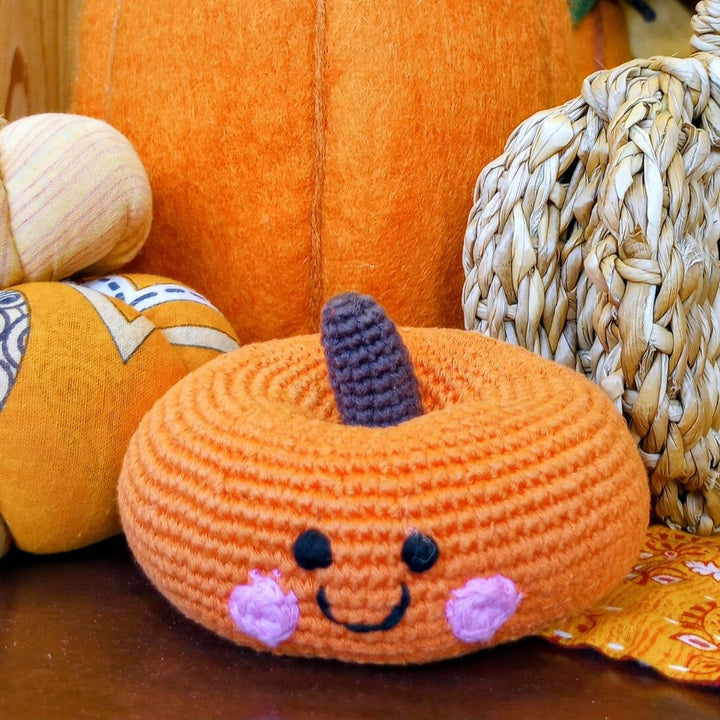 Crocheted pumpkin baby rattle with a face surrounded by real pumpkins on a wooden surface