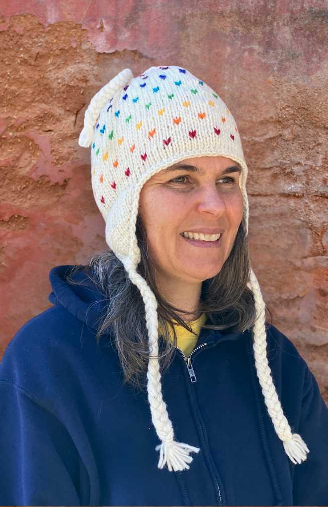 Person wearing a colorful knit hat with heart patterns against a textured wall.