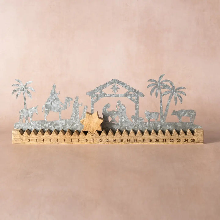 Galvanized metal nativity scene silhouette decoration with a wooden ruler base showing countdown to Christmas, set against a solid beige background.