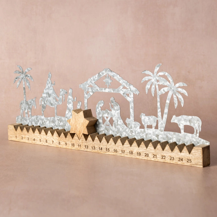 A silver metal nativity scene with silhouettes of animals, palm trees, and a stable, mounted on a wooden base printed with a 25-day countdown calendar from 1 to 25, set against a solid dusty pink background.