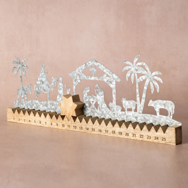 A silver metal nativity scene with silhouettes of animals, palm trees, and a stable, mounted on a wooden base printed with a 25-day countdown calendar from 1 to 25, set against a solid dusty pink background.