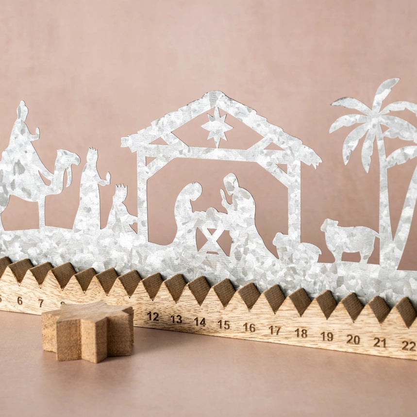 Galvanized steel Christmas nativity scene wall decor with a wooden 22-day countdown calendar base, shown against a solid dusty pink background.