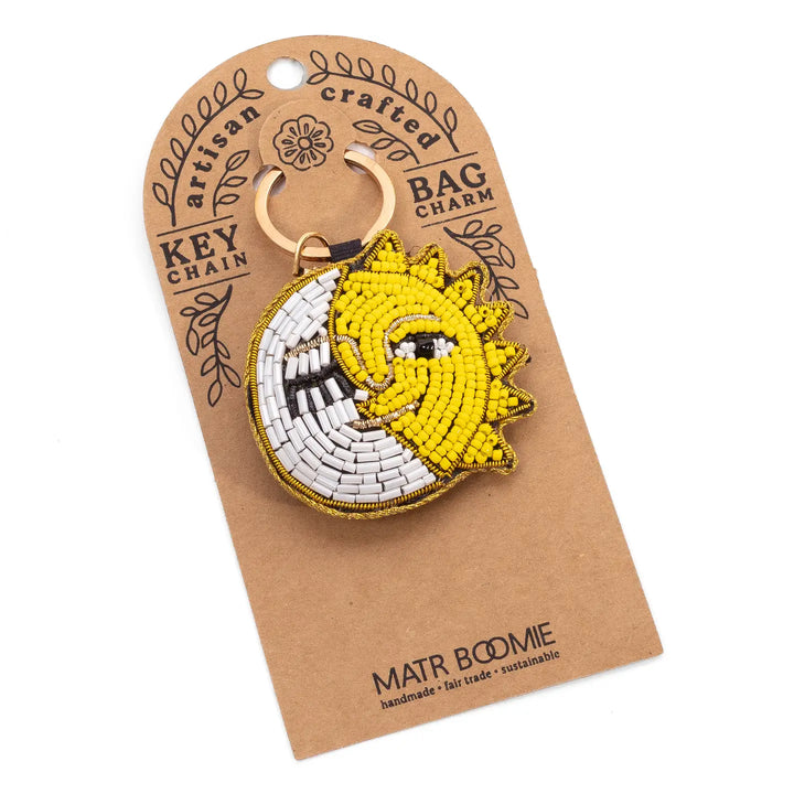 A handmade beaded keychain charm featuring a yellow sun and white crescent moon with black details, attached to a brown kraft paper display card by the brand Matr Bodmie.