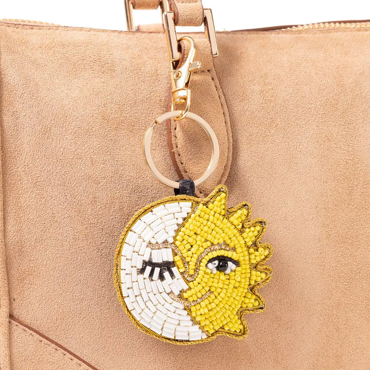 A close-up of a beige suede handbag with a gold-tone clasp and a circular beaded charm featuring a split sun and moon design in yellow and white.