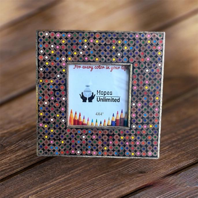 A square picture frame made from multicolored pencils, with a 4x4 photo placeholder in the center, on a wooden surface.