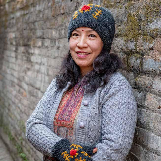 Woman wearing a gray knit outfit with embroidered gloves and hat against a stone wall.