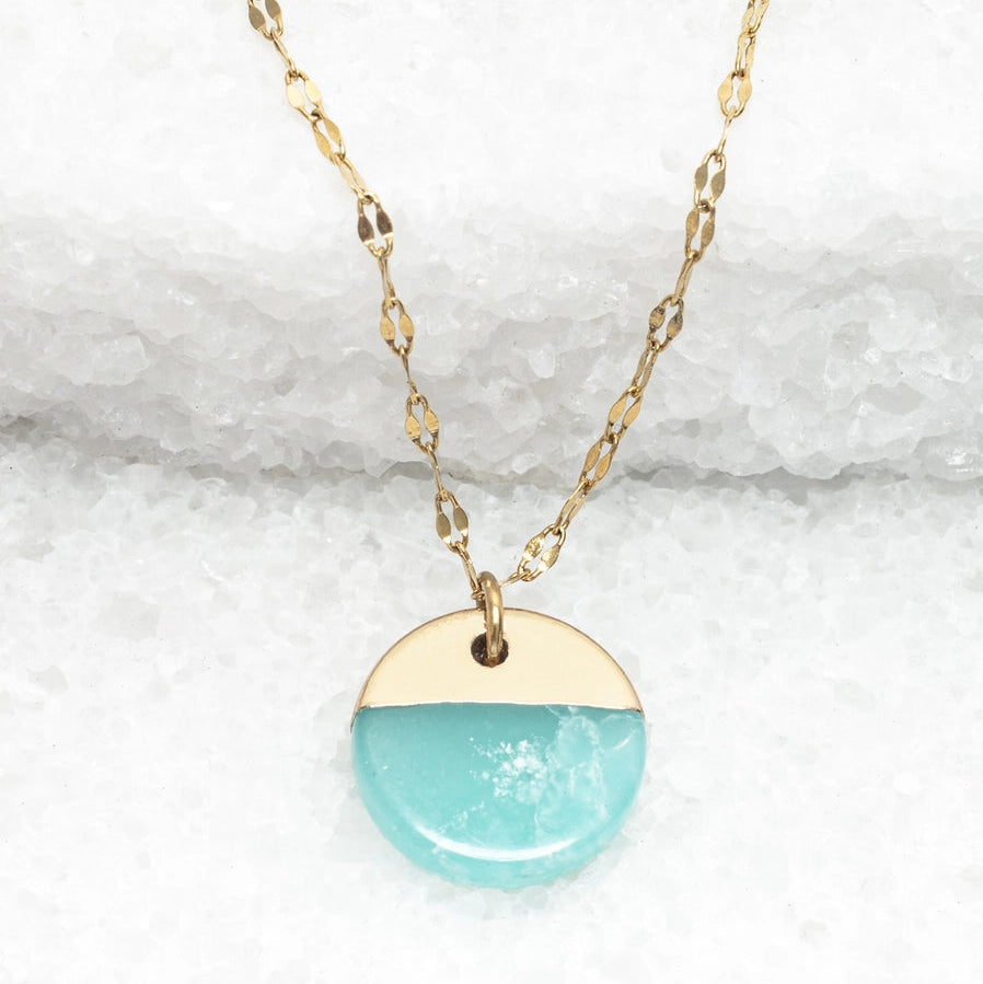A gold necklace with a round aqua pendant on a white stone background