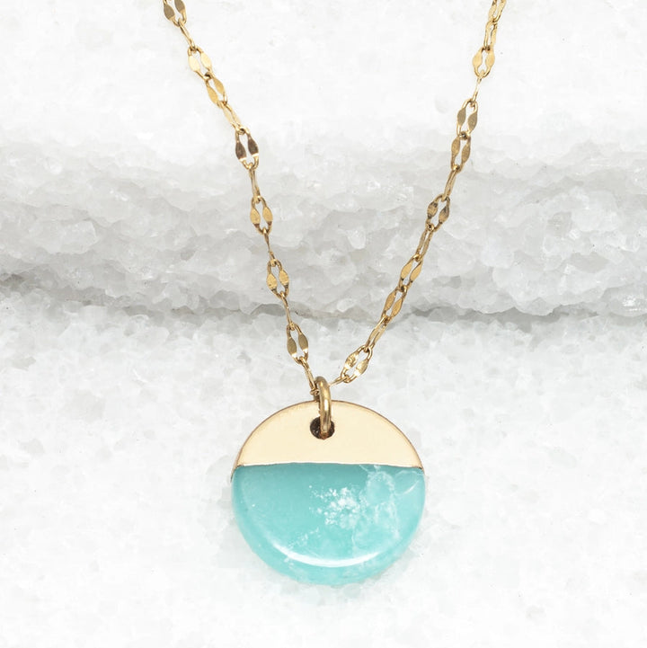 A gold necklace with a round aqua pendant on a white stone background
