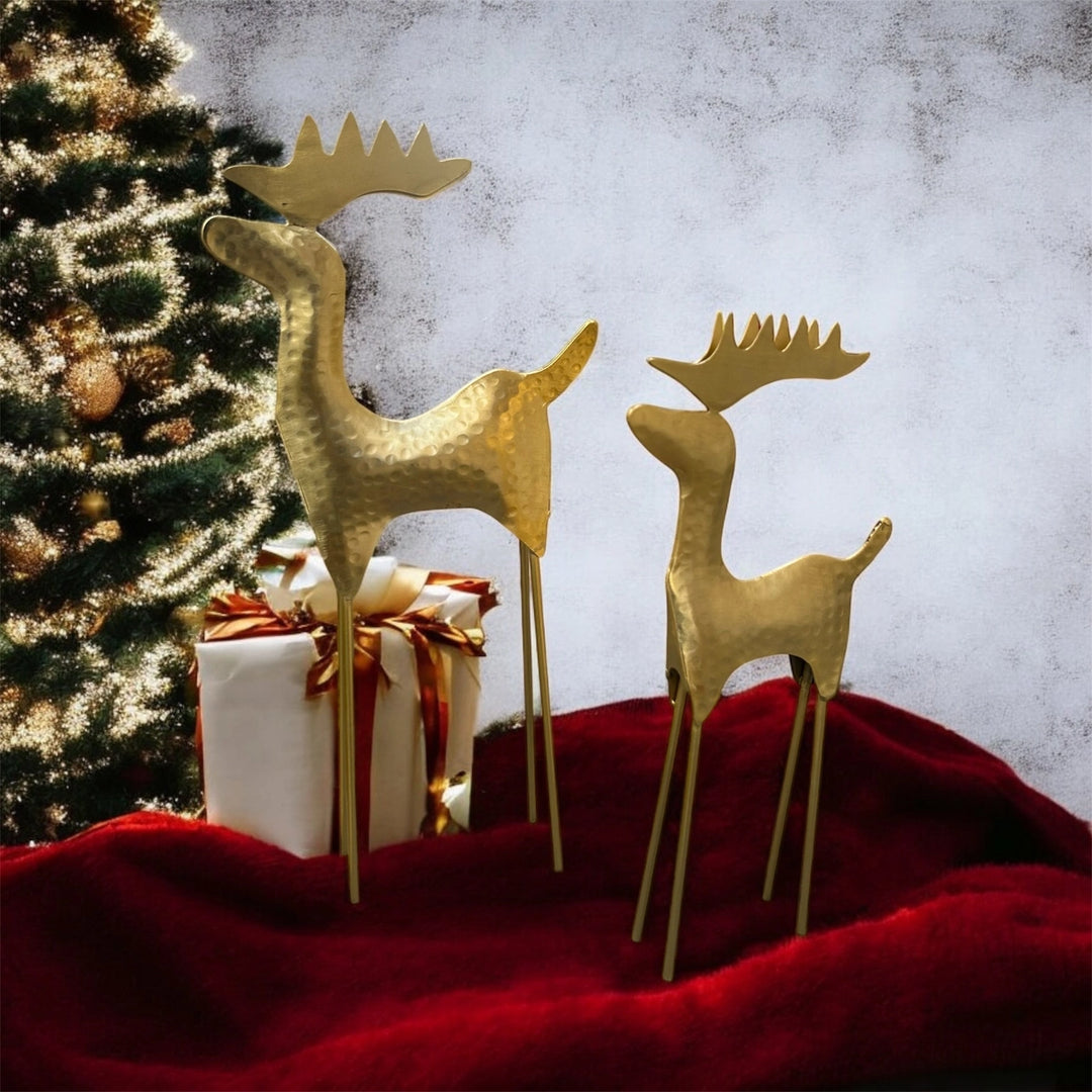 Two gold metal reindeer yard decorations with textured hammered finishes and long thin legs, displayed on a red fabric surface next to a wrapped gift box and a decorated Christmas tree against a mottled gray background.