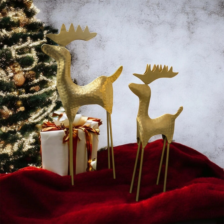 Two gold metal reindeer yard decorations with textured hammered finishes and long thin legs, displayed on a red fabric surface next to a wrapped gift box and a decorated Christmas tree against a mottled gray background.
