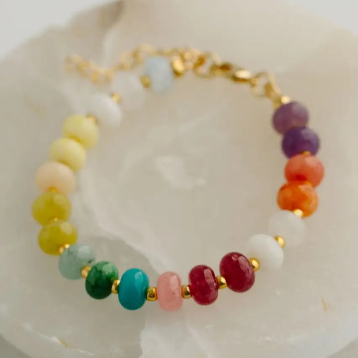 A beaded bracelet with faceted jade beads in a rainbow sequence, separated by small gold spacer beads and connected by a gold clasp, displayed on a pale stone surface.