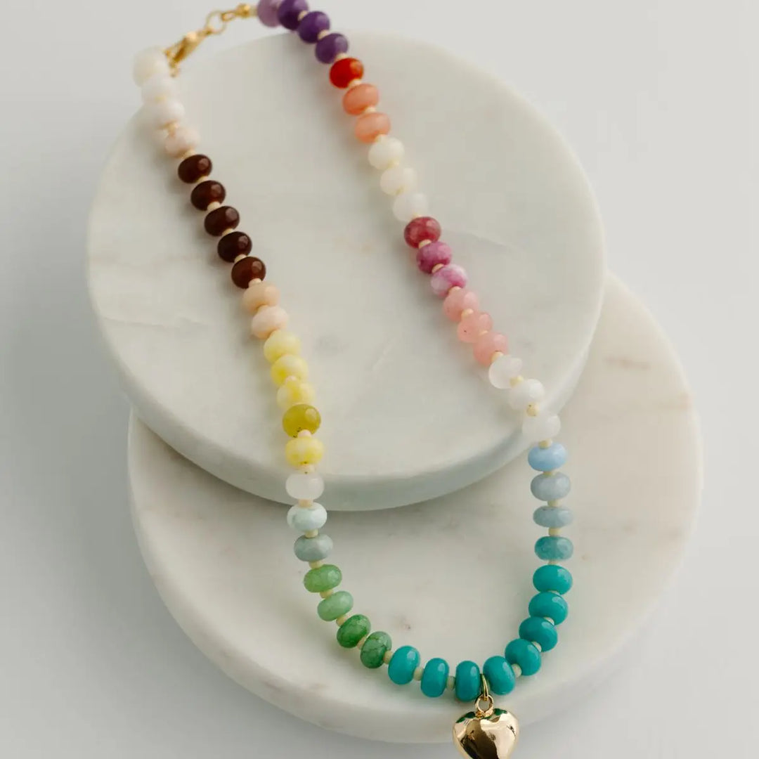 A beaded rainbow necklace with a gold heart charm, displayed on stacked white marble round trays against a light gray background.