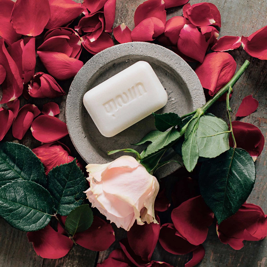 Hand-crafted rose scented bar of soap on soap dish surrounded by red rose petals with a pretty pink rose.