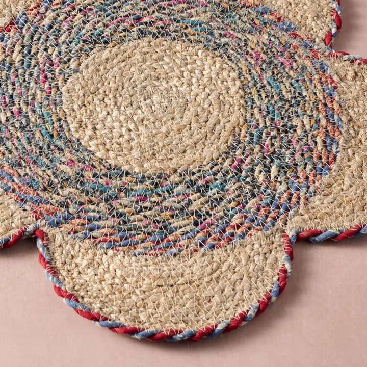 A close-up of a round braided placemat with a flower-like silhouette, featuring a natural beige center and concentric rings of blue, pink, and red braided accents on a dusty pink background.