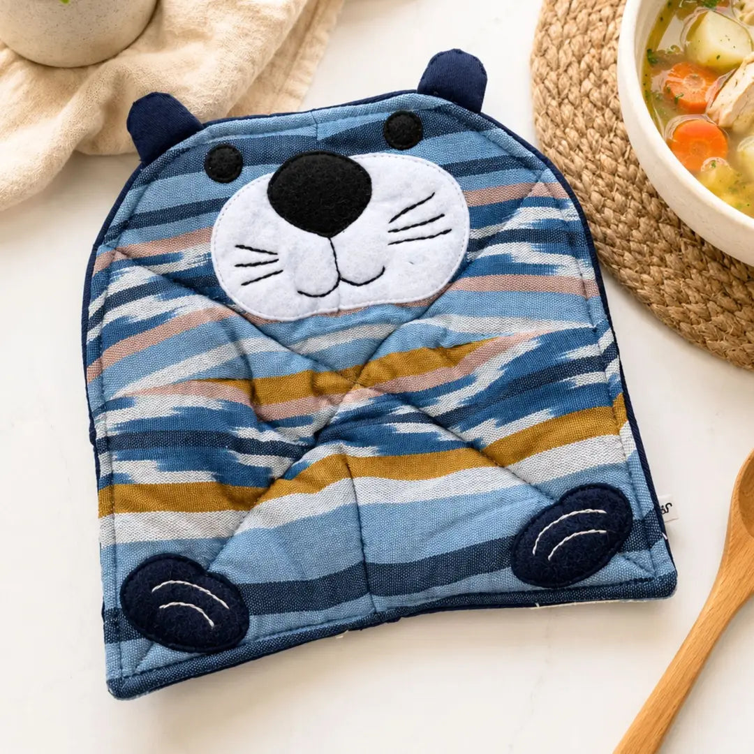 A quilted pot holder shaped like a cartoon otter face, featuring blue, navy, white, beige, and mustard yellow stripes, styled on a white surface with a bowl of soup and a wooden spoon nearby.