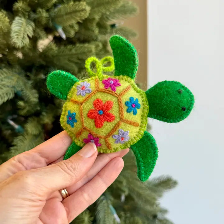 A handmade felt turtle ornament with a yellow-green body, green flippers and shell accents, and colorful embroidered flowers, held in a person's hand in front of a Christmas tree.