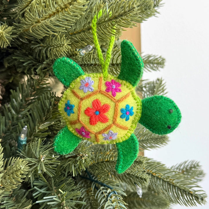 A handmade felt turtle Christmas ornament hanging on a green pine Christmas tree. The ornament features a light green body with embroidered colorful flowers, green felt legs and head, and a green hanging loop.