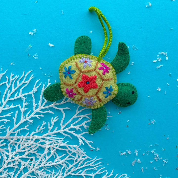 A handmade felt sea turtle ornament with a light green shell decorated with colorful embroidered flowers, green felt limbs, and a green hanging loop, displayed on a bright blue background with white coral-like decoration.