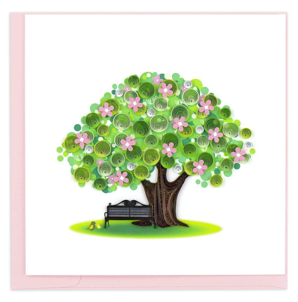 A handmade greeting card featuring a quilled design of a tree with pink flowers and a bench, on a green background.