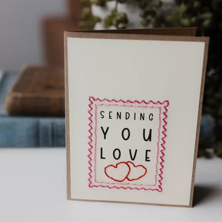 A square greeting card with a kraft brown border and an off-white center, featuring the words 'SENDING YOU LOVE' in black with a pink stitched border and two red hearts at the bottom.