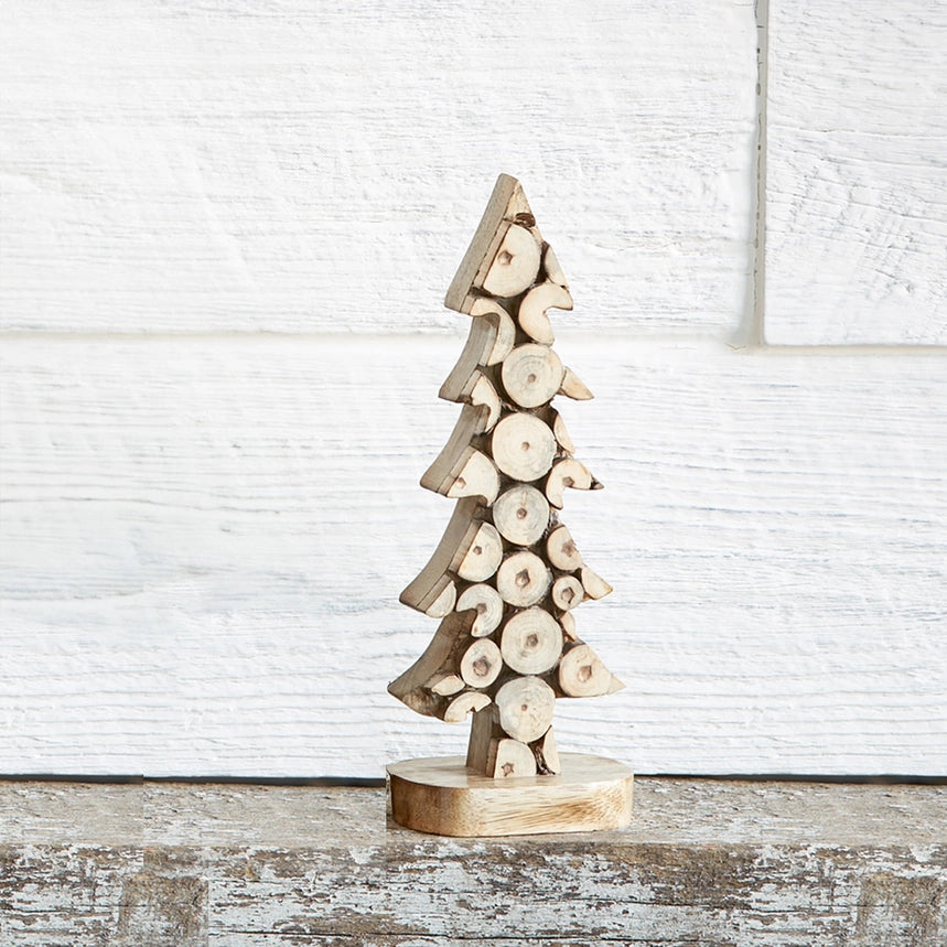 A small decorative Christmas tree made from stacked natural wood logs on a round wooden base, set against a white paneled wall and rustic wooden surface.