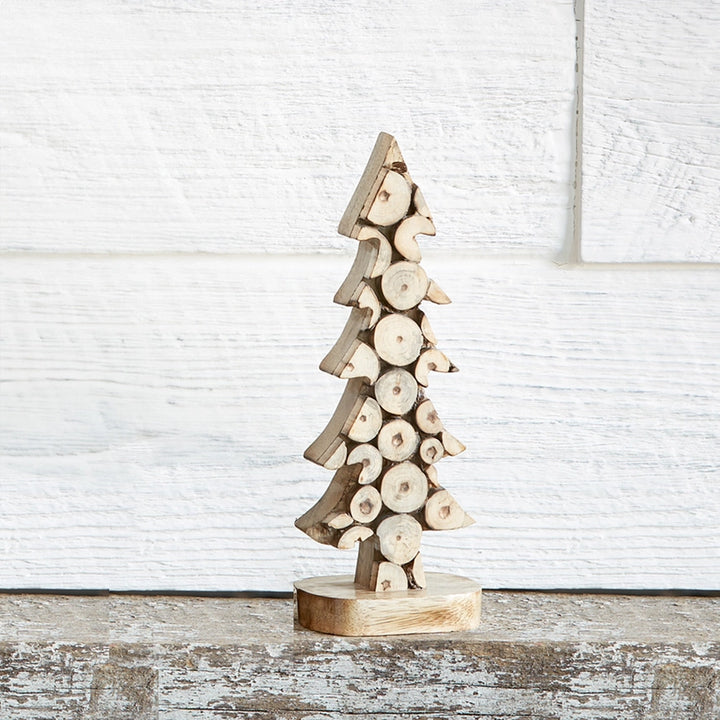 A small decorative Christmas tree made from stacked natural wood logs on a round wooden base, set against a white paneled wall and rustic wooden surface.