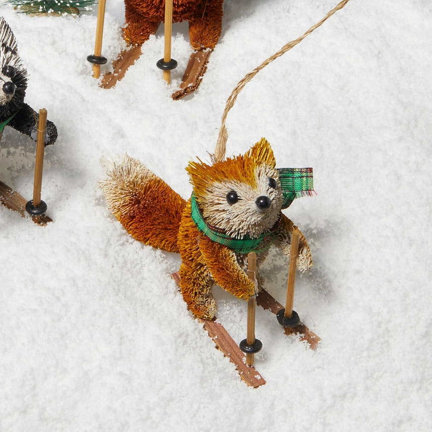 A small buri fox ornament wearing a green scarf and plaid hat, posed as if skiing in white snow, with wooden skis and a hanging string.