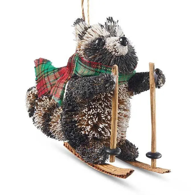 A raccoon-shaped hanging ornament with a red and green plaid scarf and a pair of wooden skis, shown against a plain white background.