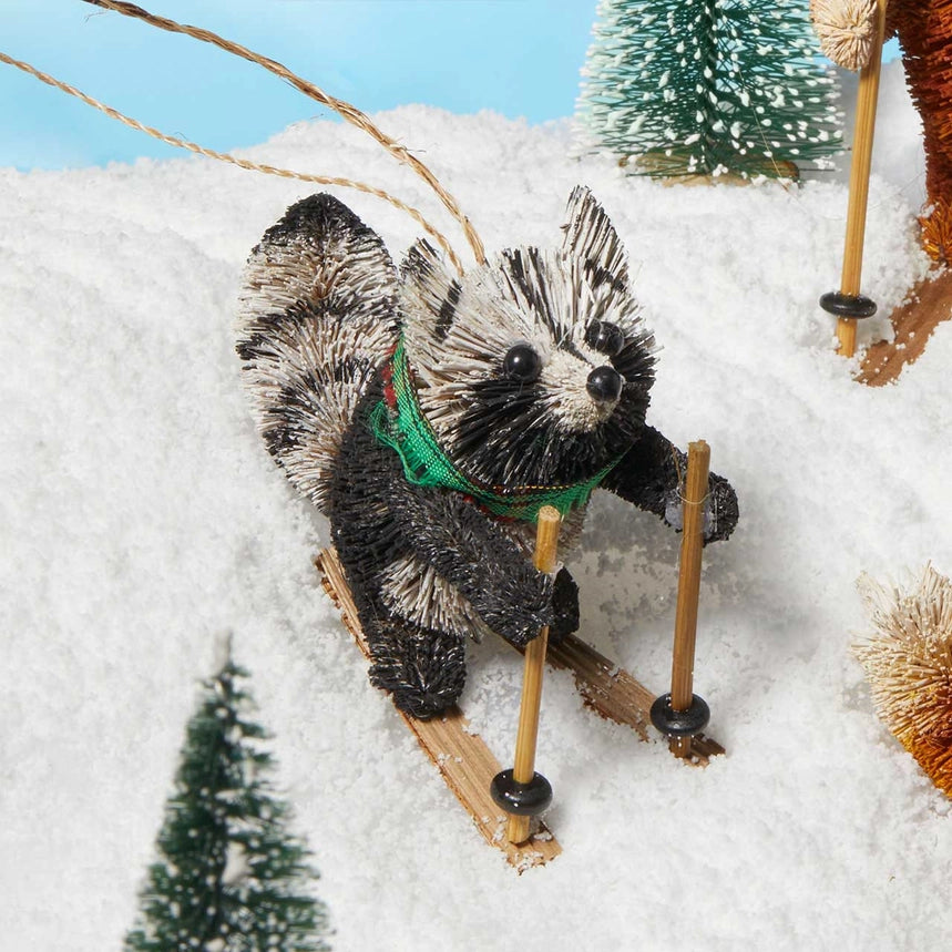 A decorative raccoon figurine wearing a green harness and skiing with small wooden poles, set in a snowy miniature scene with small Christmas trees.
