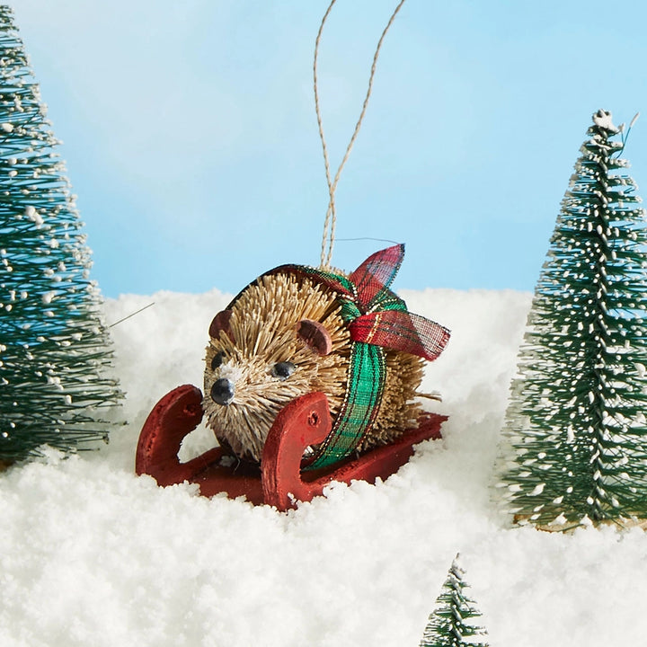 A decorative hedgehog ornament sitting in a red sled, set in white faux snow with miniature green trees and a blue sky backdrop.