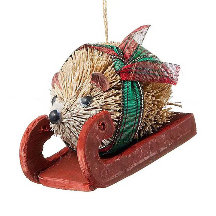 A Christmas hedgehog ornament sitting in a red wooden sled, decorated with a green and red plaid ribbon bow, on a white background.