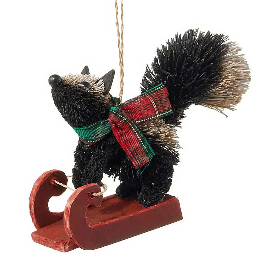 A black bristle skunk ornament wearing a red and green plaid bow and standing on a red wooden sleigh, suspended by a string against a white background.