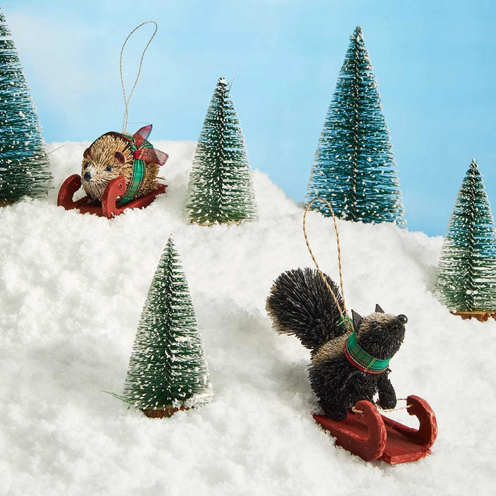 Two Christmas tree-sitter ornaments shaped like a hedgehog and skunk in red sleds, displayed in a snowy winter scene with miniature pine trees against a light blue sky backdrop.