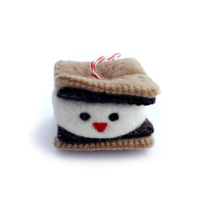 A handmade wool ornament resembling a s'more with a smiling face, in shades of beige, brown, and black, with a red ribbon on top.