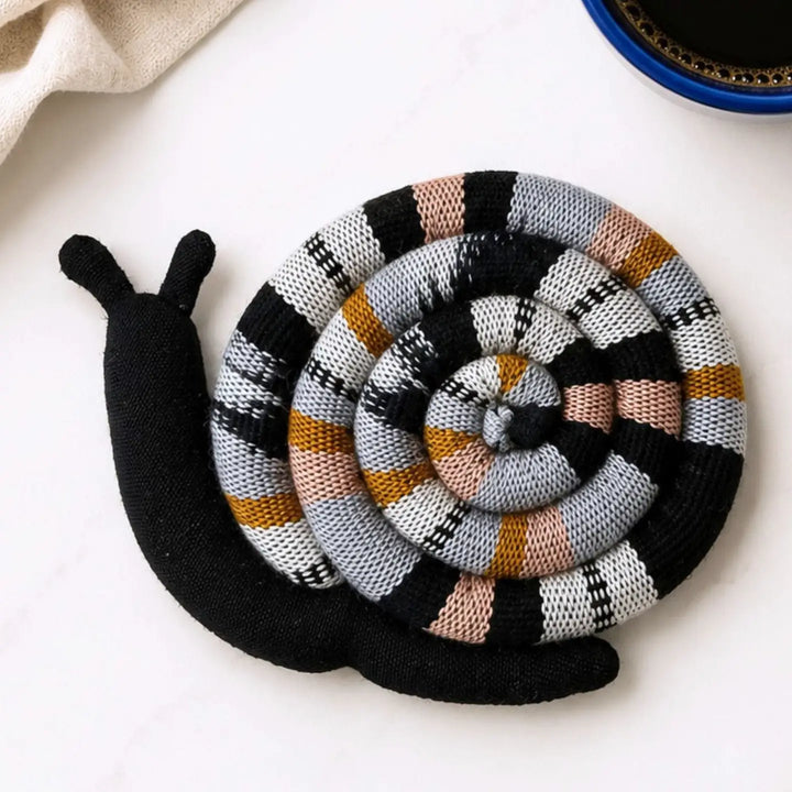 A knitted snail-shaped coaster or trivet with a black body and a spiral shell pattern in black, white, gray, peach, and mustard, photographed on a light surface with partial props at the edges.