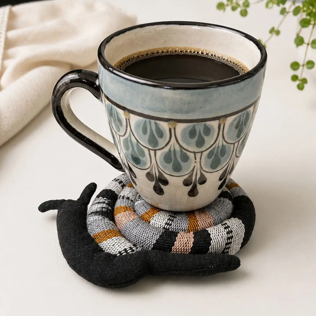 A ceramic coffee mug with a blue-gray and cream teardrop pattern sits on a black plush mouse-shaped coaster over a striped knit coaster pad, on a light tabletop with a cream cloth and green leaves in the background.
