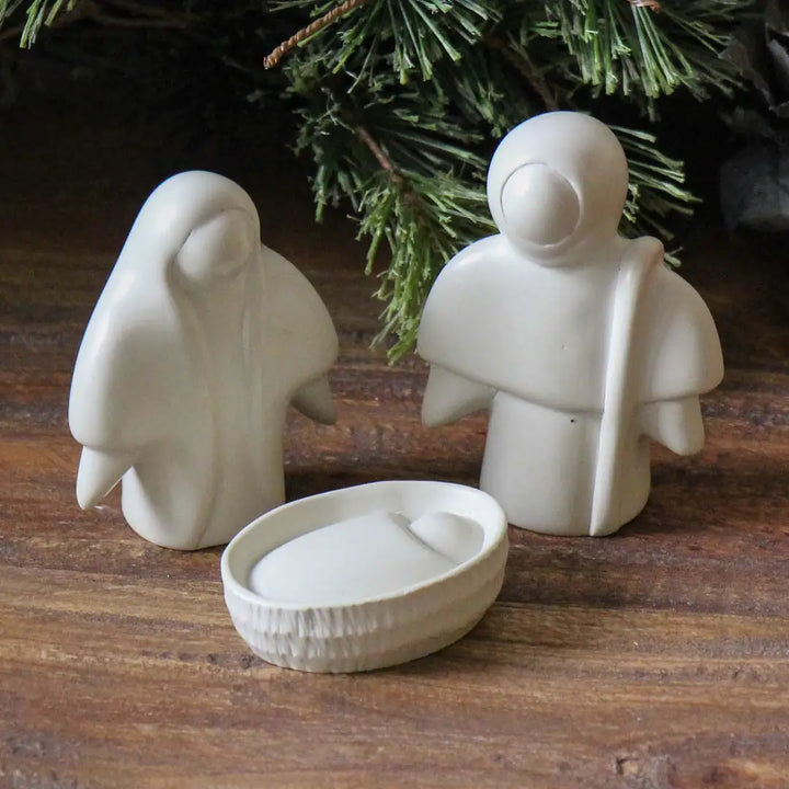 Three minimalist white soapstone figurines arranged on a wooden surface with green pine branches in the background. The set includes a Mary figure on the left, a Joseph figure holding a staff on the right, and a shallow oval Baby Jesus in the foreground.
