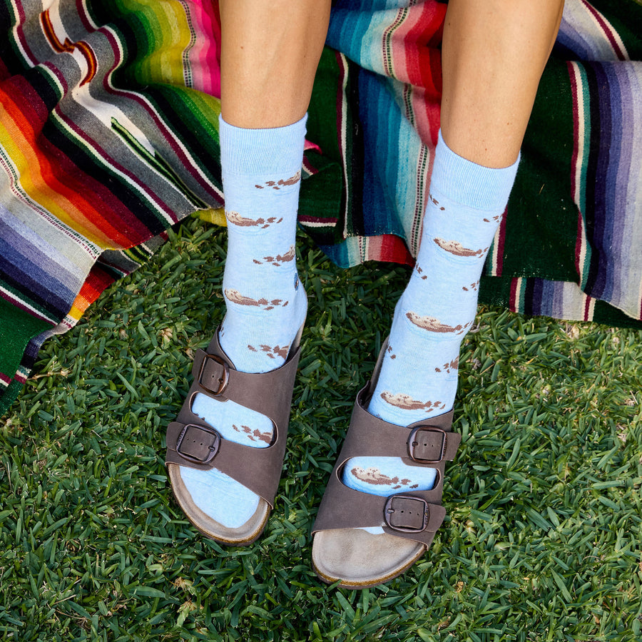 Person wearing blue socks with sea otter pattern and brown sandals on grass, colorful blanket in background