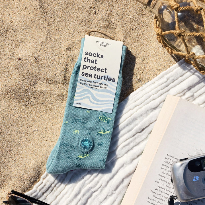 A single light blue sock with a printed sea turtle pattern and a decorative button, laid on a white blanket on a sandy beach. The sock has a paper tag that reads 'conscious sock socks that protect sea turtles'.
