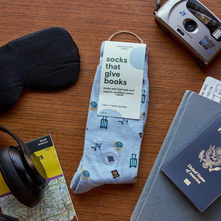 A pair of light blue crew socks with a small airplane and book pattern, featuring a white paper tag that reads 'socks that give books' and 'Christian's Shop'. The socks are styled in a flat-lay on a wooden table surrounded by travel-themed props including a passport, headphones, a camera, and a sleep mask.