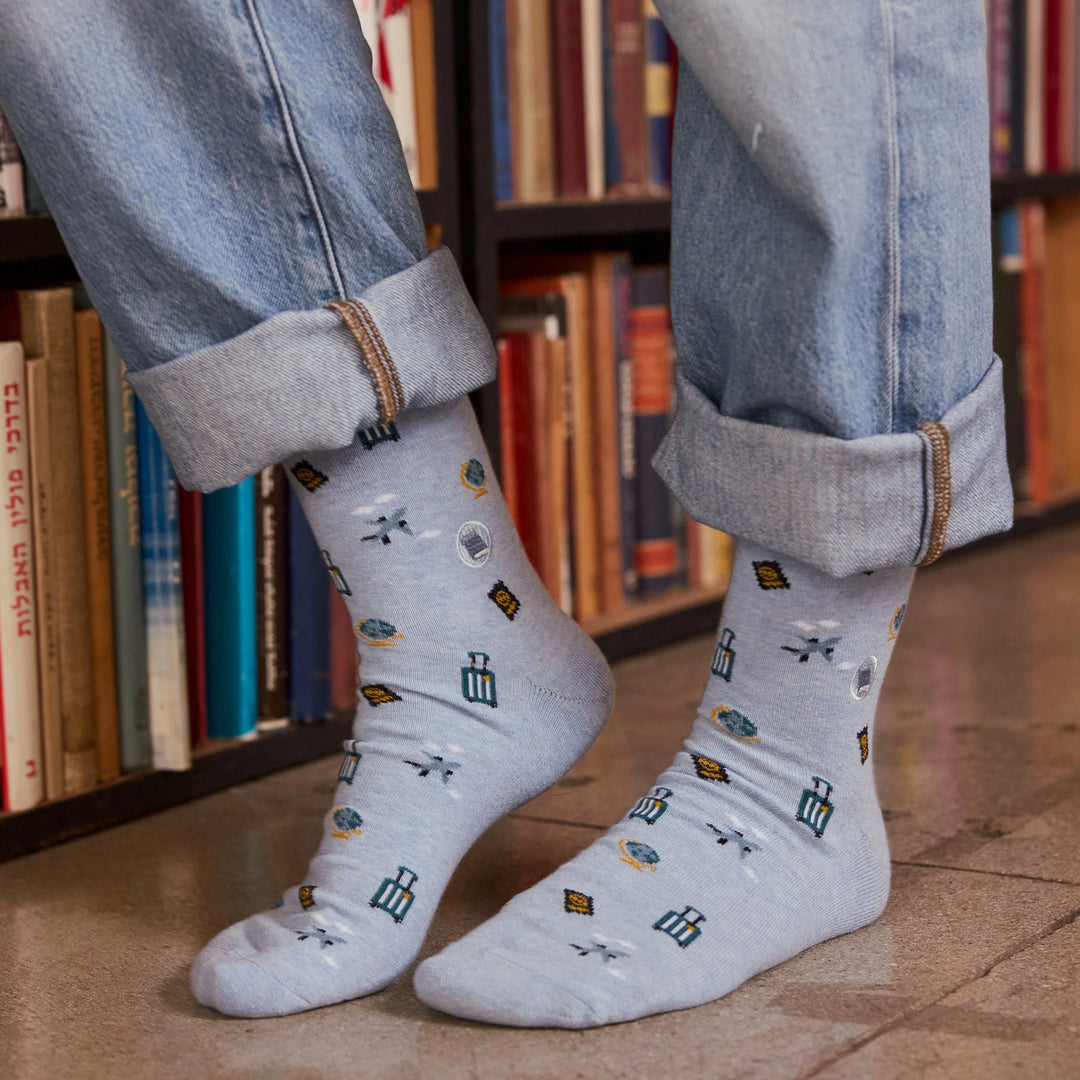 A person wearing light gray crew socks with a pattern of small blue, yellow, and white icons, paired with light blue jeans cuffed at the ankles, standing in front of a bookshelf.