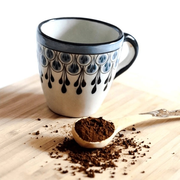Ceramic mug with decorative black patterns on a wooden surface with coffee grounds and a spoon.