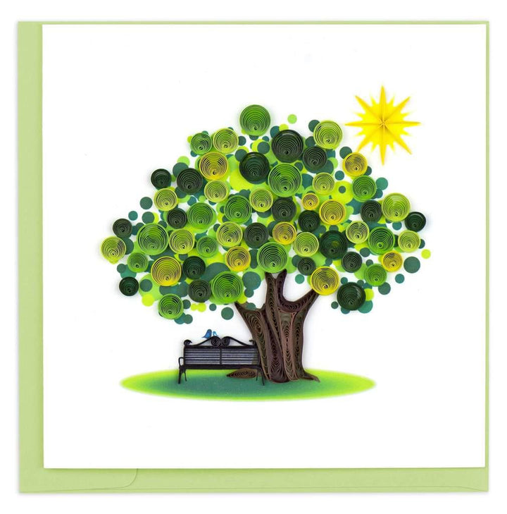 A greeting card featuring a quilled design of a tree with green leaves and a sun, beside a bench, crafted from paper strips.