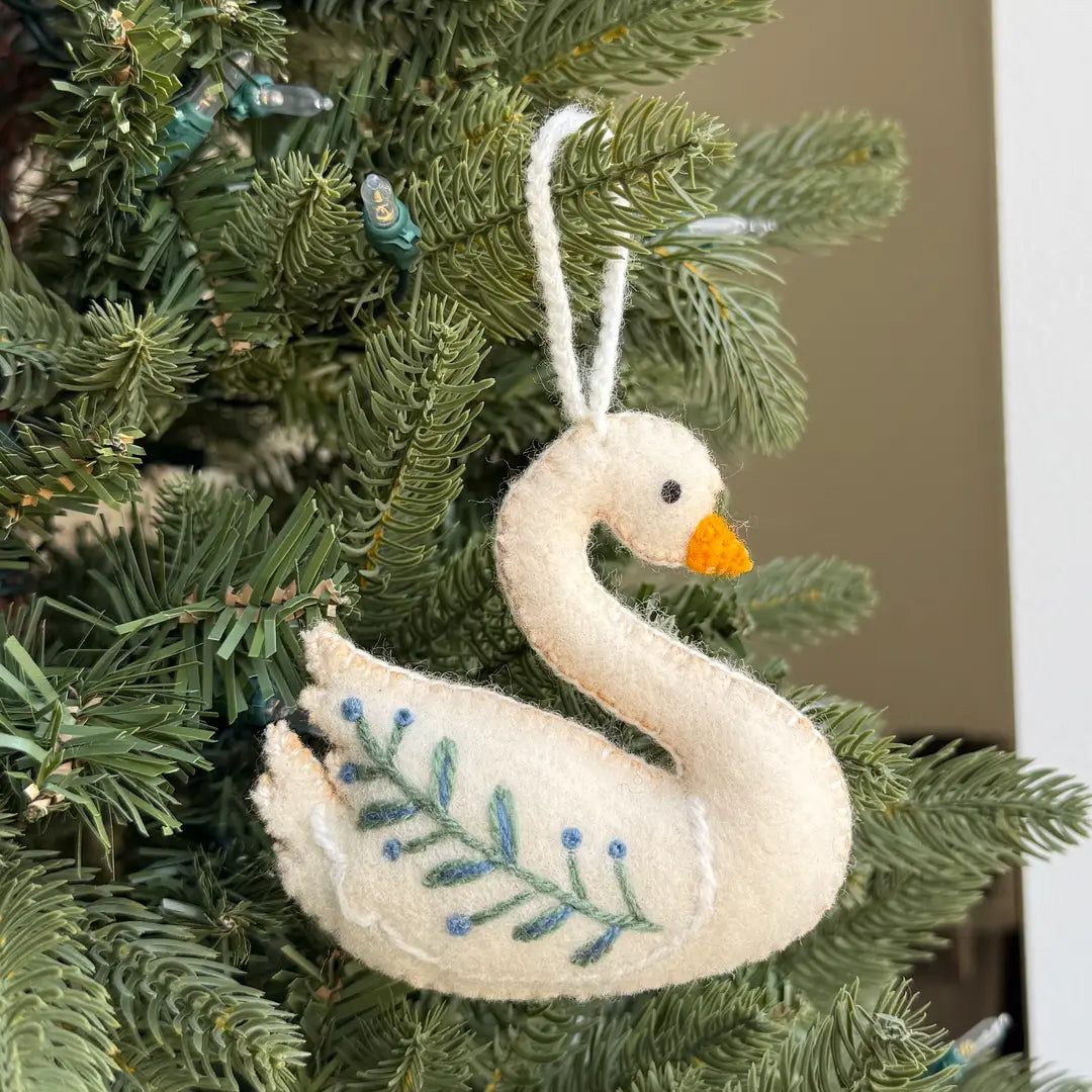 A felt swan ornament with a white body, orange beak, and blue embroidered leaves, hanging on a green Christmas tree.