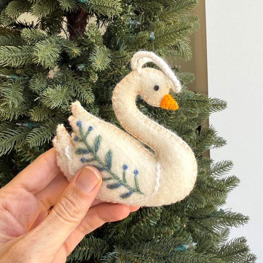 A hand holding a white felt swan ornament with blue embroidered leaves and berries, set against a green Christmas tree background.