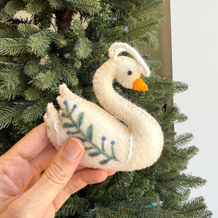 A hand holding a white felt swan ornament with blue embroidered leaves and berries, set against a green Christmas tree background.